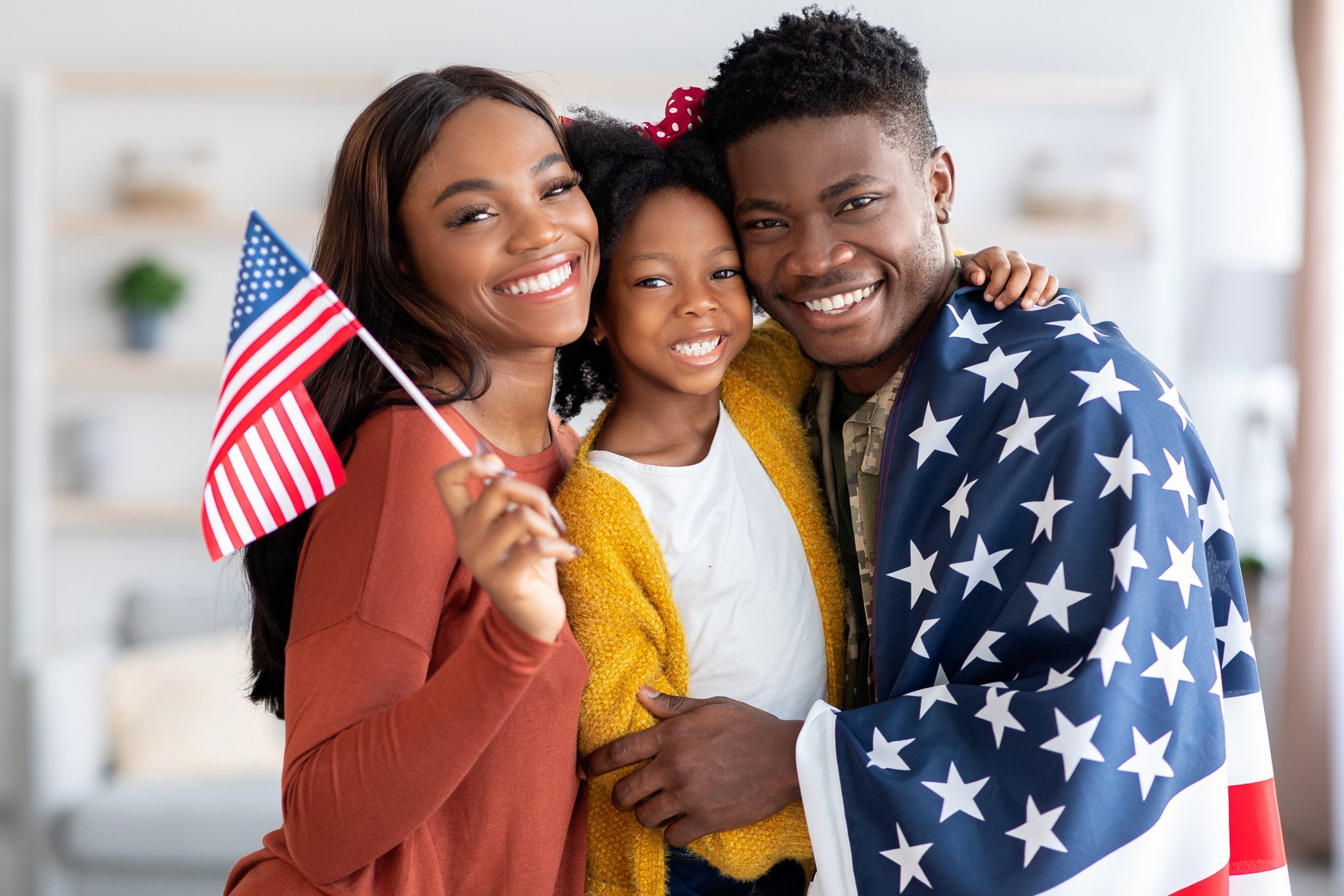 Happy black military man wrapped in american flag posing with wife and little daughter at home after returning from army, cheerful family celebrating reunion, embracing and smiling at camera